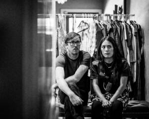 A black-and-white photo of Joann Mercado Hicks seated beside Byron Watkins, framed by a rack of wardrobe pieces as they discuss film production plans.