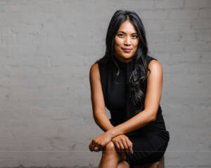 Joann Mercado Hicks seated against a brick wall in a sleek black dress, looking poised and confident.