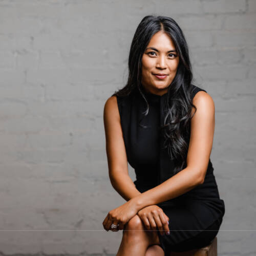 Joann Mercado Hicks seated against a brick wall in a sleek black dress, looking poised and confident.