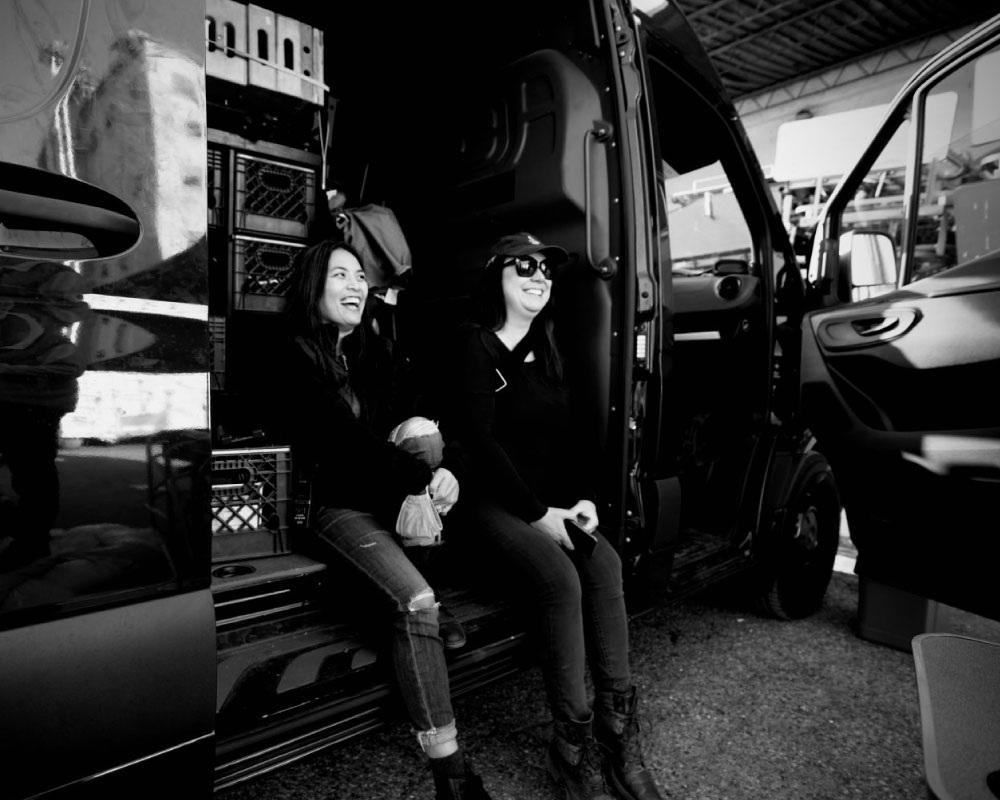 A candid black-and-white shot of Joann Mercado Hicks and Sara Bourbeau relaxing in the open side door of a production van, sharing a light moment on set.