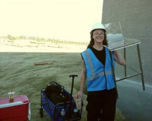 Abby Edwards, Production Assistant, in a blue safety vest and white hard hat smiles while carrying a clear acrylic chair at an outdoor construction site. She holds an iced drink and stands next to a wagon loaded with supplies.