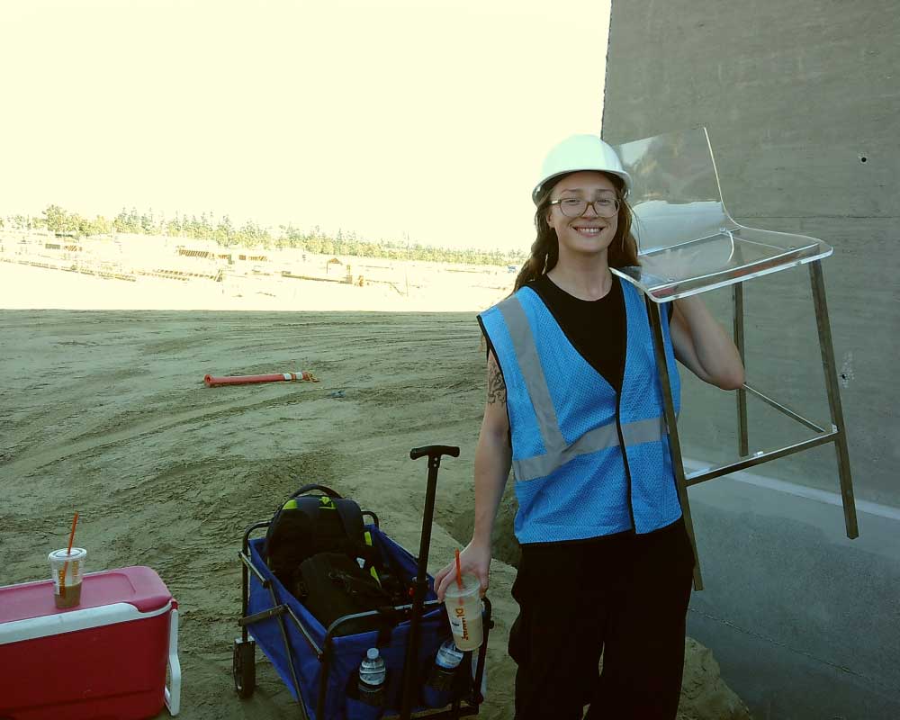 Abby Edwards, Production Assistant, in a blue safety vest and white hard hat smiles while carrying a clear acrylic chair at an outdoor construction site. She holds an iced drink and stands next to a wagon loaded with supplies.