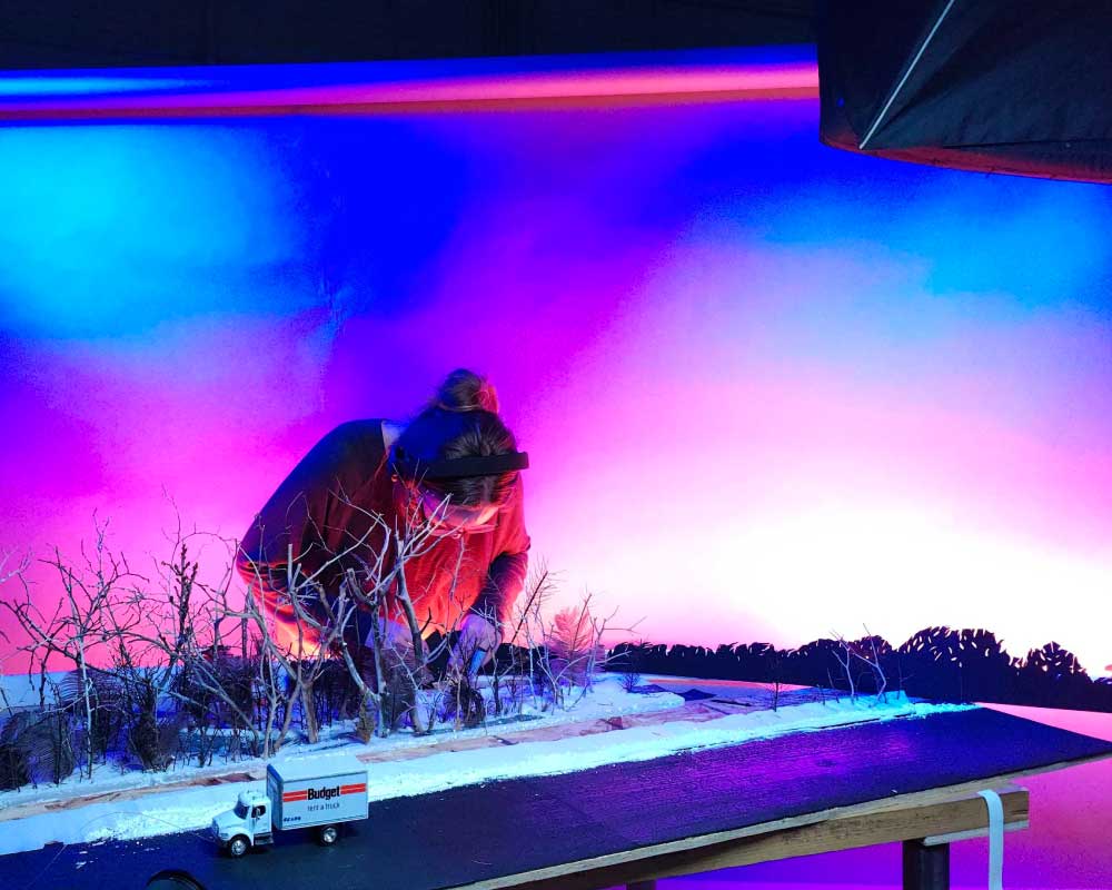 Abby Edwards, Production Assistant, wearing a headlamp, leans over a detailed miniature set of a winter landscape, adjusting small trees and snow under vibrant pink and blue lighting in a studio setting.