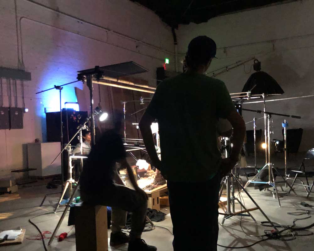 A dimly lit film production set filled with lighting rigs and equipment. Abby Edwards, Production Assistant, stands silhouetted in the foreground while another crew member works near a detailed miniature set under studio lights.