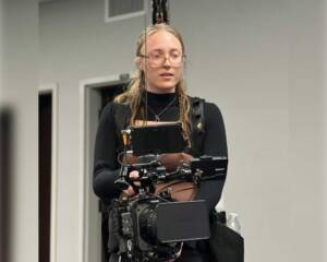 Abby Edwards, Production Assistant, operates a professional Sony camera rig with a stabilizing vest in an indoor filming environment, focused on capturing the perfect shot.