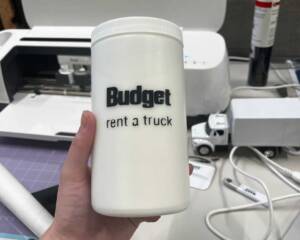A close-up of Abby Edwards, Production Assistant, holding a white cylindrical container with a custom-made "Budget Rent a Truck" label, with a workspace full of crafting tools and materials in the background.