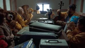 Windsong Productions cast and crew dressed in yellow costumes and puppets performing as the Kenosha Kicker Polka Band in a reimagined Home Alone scene inside a van filled with luggage