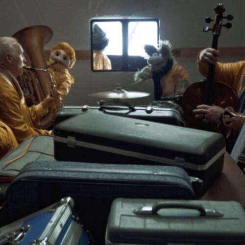 Windsong Productions cast and crew dressed in yellow costumes and puppets performing as the Kenosha Kicker Polka Band in a reimagined Home Alone scene inside a van filled with luggage