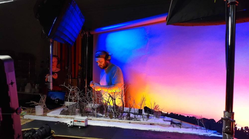 Behind-the-scenes view of a Windsong Productions crew member setting up a handcrafted winter miniature set with a sunset-colored backdrop for the Home Alone remake