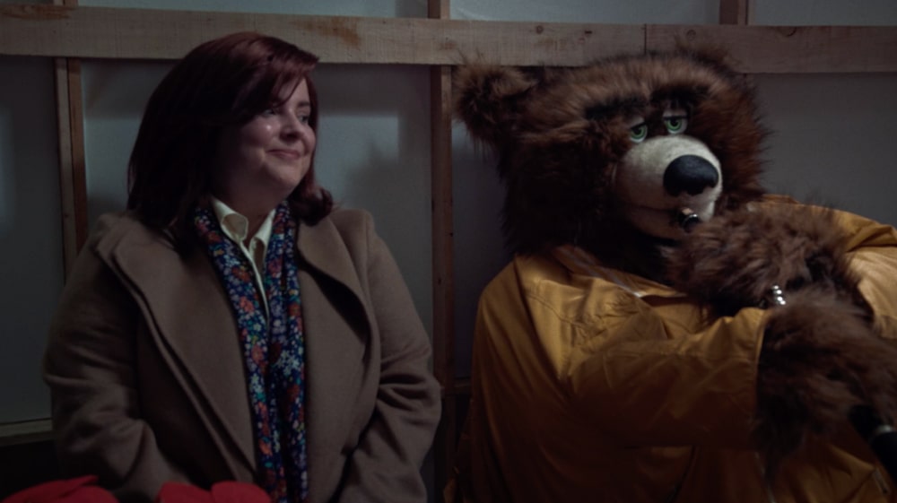 Close-up of actress Chelsea Gue as Kevin’s mom sitting beside a bear-costumed puppet playing a clarinet in Windsong’s Home Alone re-creation