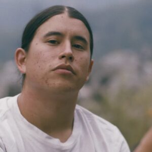 Serious young man with long hair wearing a white t-shirt, gazing into the camera with a natural outdoor landscape in the background.
