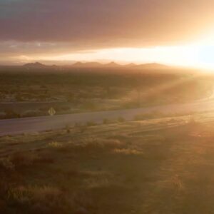 Scenic desert highway at sunset with golden sunlight casting over rolling hills and distant mountain ranges.