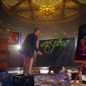 Artist spray-painting the word 'Inspire' on a large canvas under a grand theater ceiling, surrounded by paintings and creative supplies.